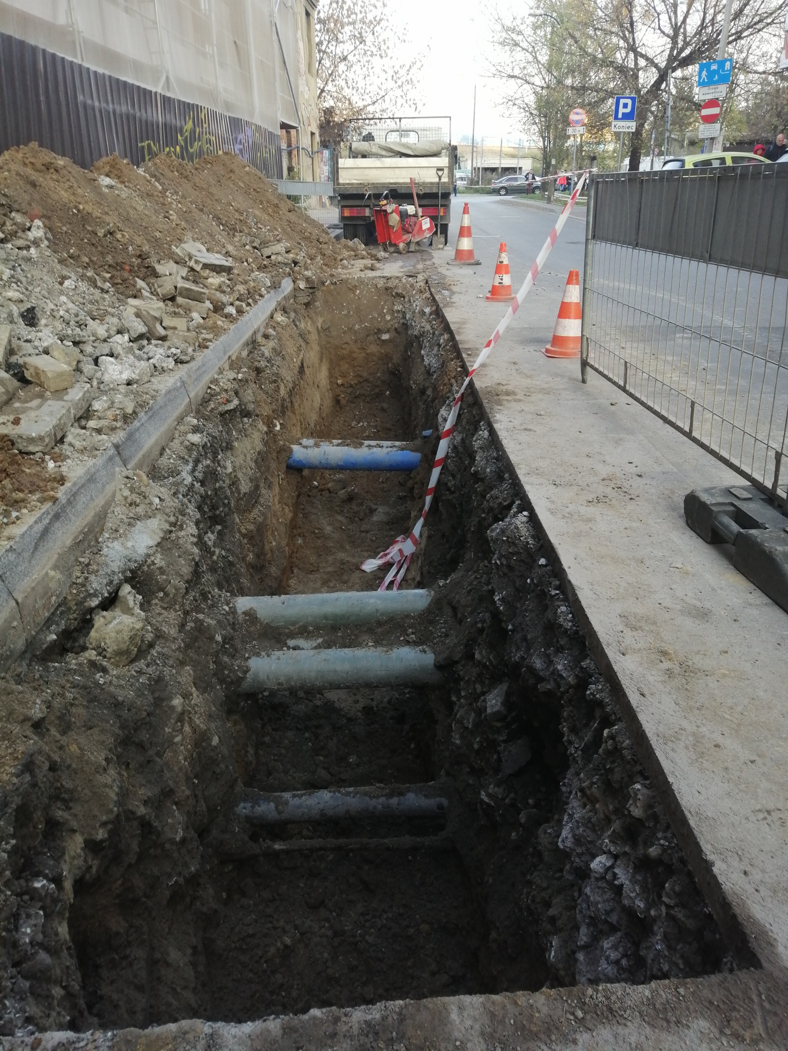 Głęboki wykop w ziemi z widocznymi rurami, zabezpieczony taśmą ostrzegawczą i pachołkami, na tle ulicy z zaparkowanymi samochodami i budynkami.