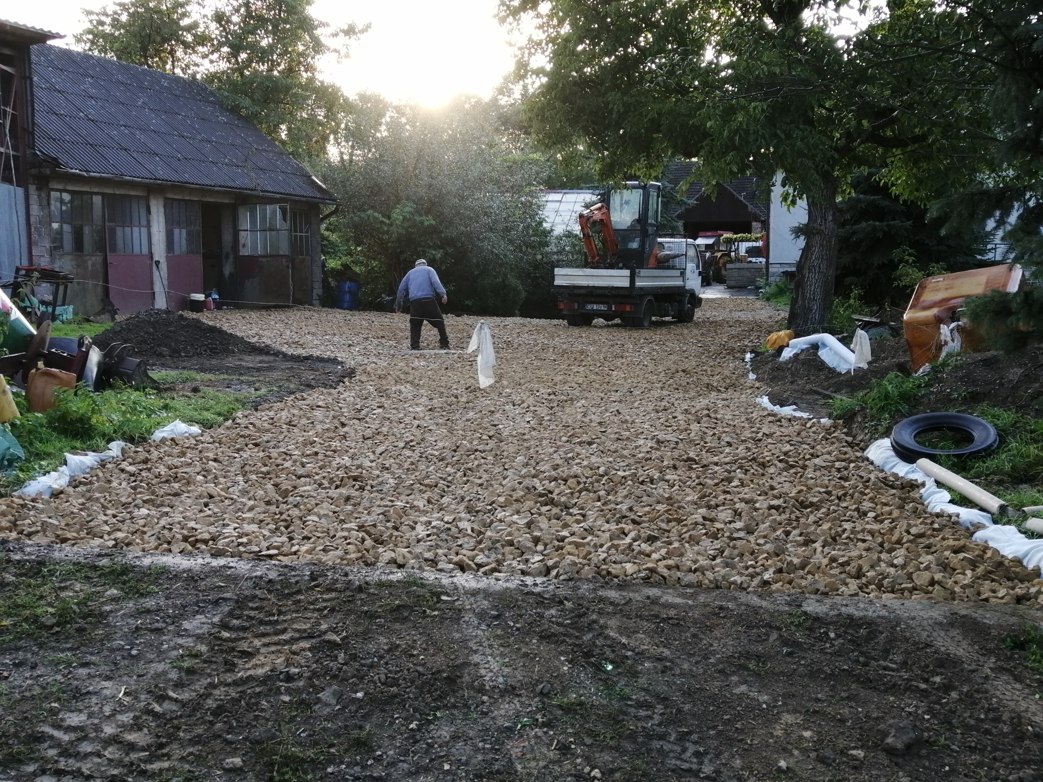 Nierówna, kamienista droga w trakcie budowy na posesji, z koparką na ciężarówce w tle i pracownikiem w niebieskiej kurtce, widok na budynek gospodarczy z lewej strony.