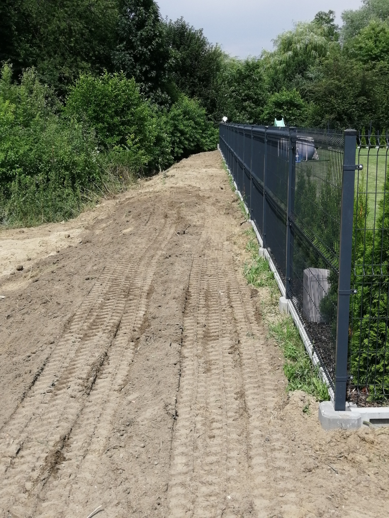 Świeżo wyrównany teren z widocznymi śladami opon maszyny budowlanej, przylegający do nowego, metalowego ogrodzenia panelowego na betonowej podmurówce, w tle zieleń drzew i krzewów.