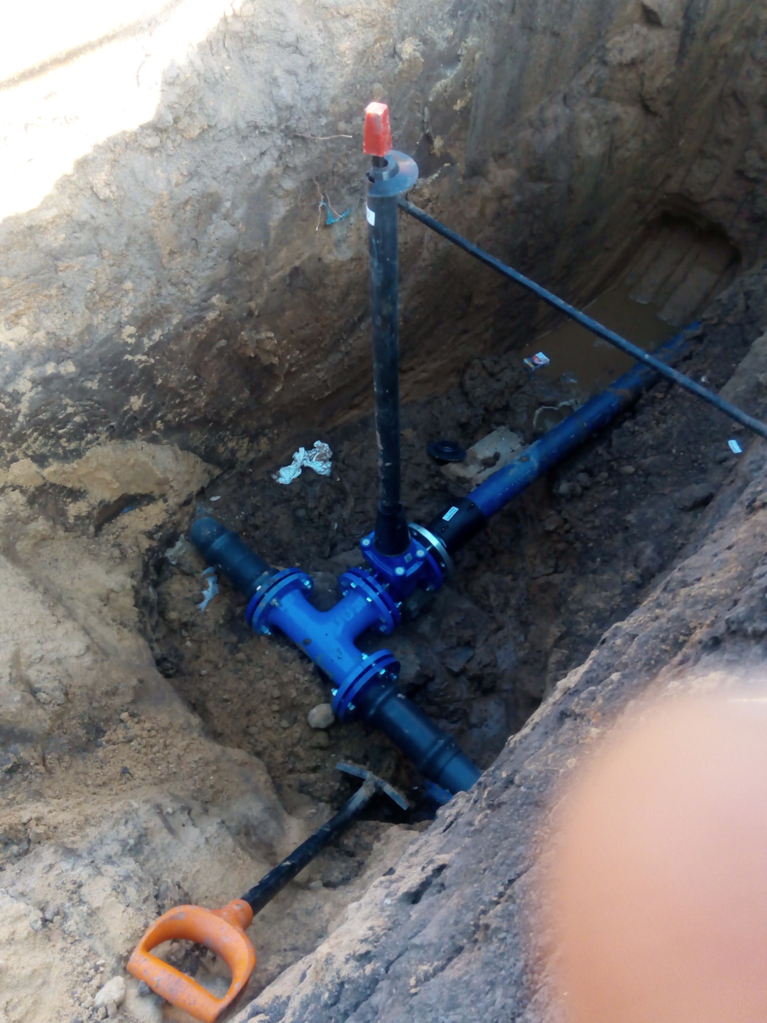 Głęboki wykop z widocznymi elementami instalacji wodno-kanalizacyjnej: niebieskie złączki i czarne rury, częściowo zanurzone w wodzie. Na dnie leży łopata z pomarańczową rączką. W tle widoczny...