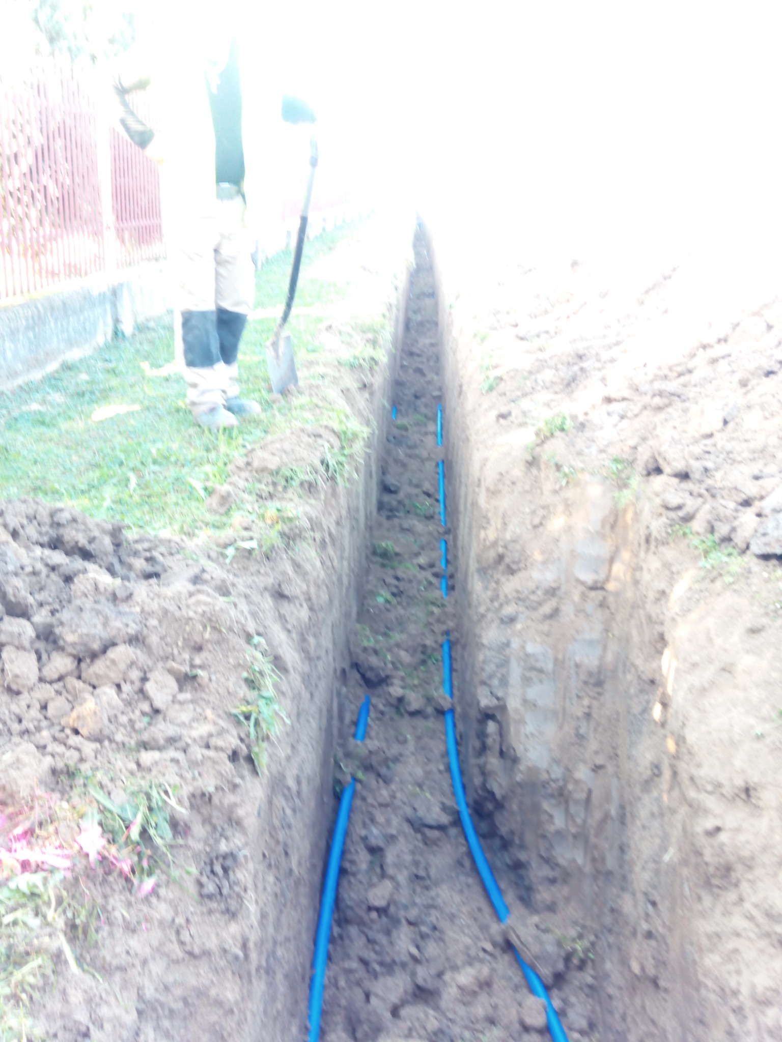 Wąski wykop w ziemi z ułożonymi niebieskimi rurami, widoczna postać z łopatą w tle, zdjęcie wykonane w słoneczny dzień.