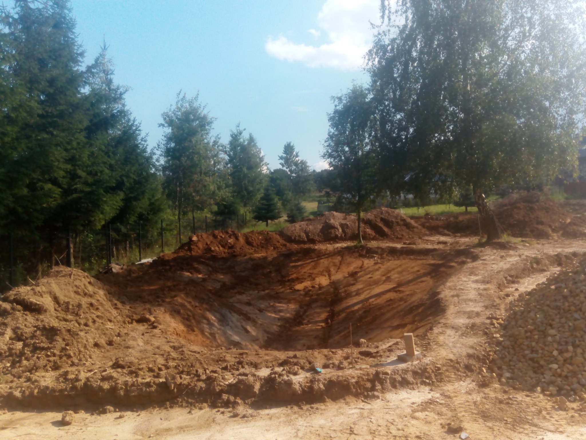 Wykopany dół w Mogilanach, otoczony stertami ziemi i drzewami, prawdopodobnie pod budowę szamba lub oczka wodnego.