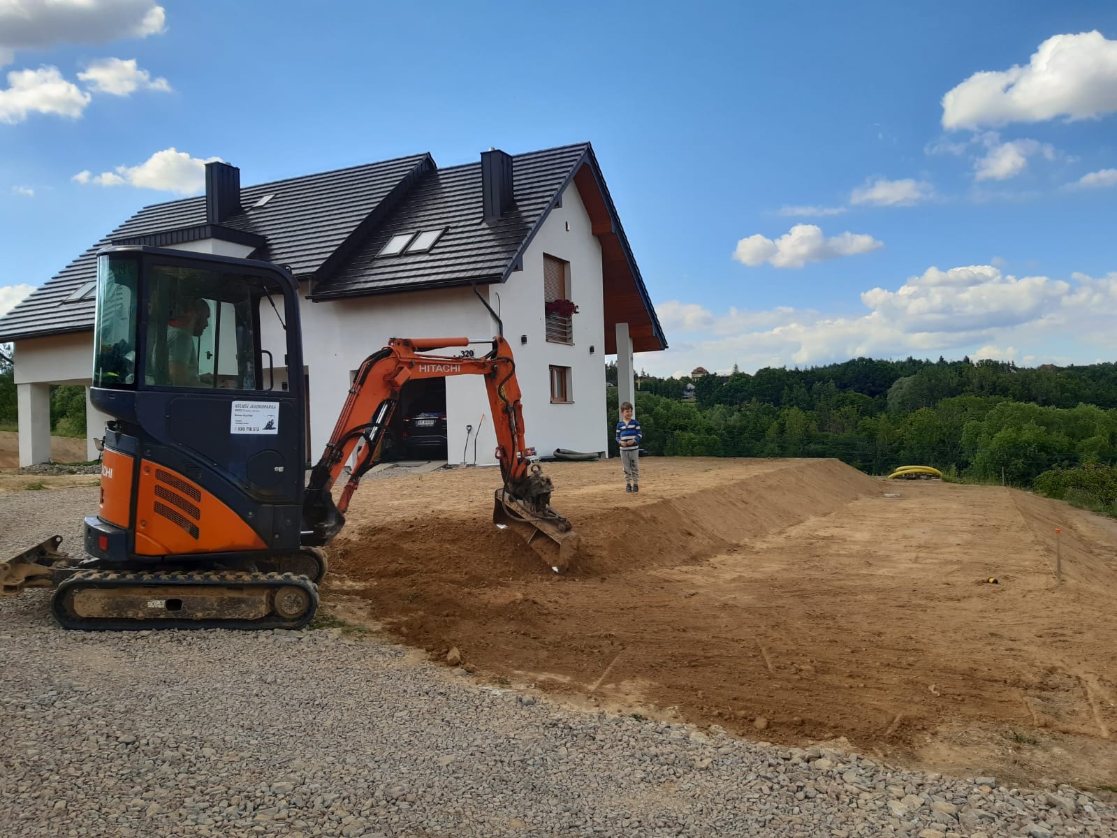 Pomarańczowo-czarna minikoparka Hitachi na żwirowym podłożu, w tle biały dom z ciemnym dachem i chłopiec stojący na wyrównanym terenie z ziemi, błękitne niebo z chmurami.