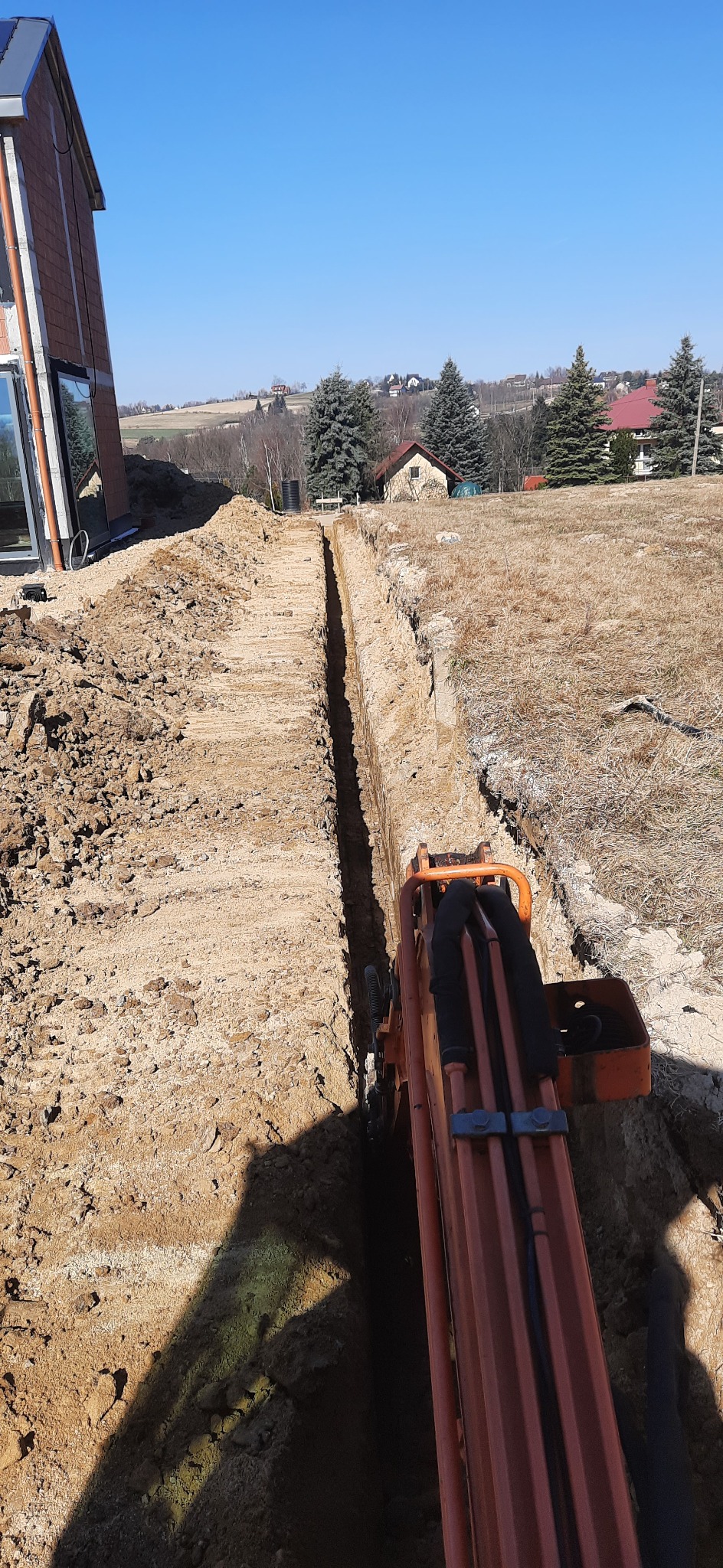 Wąski wykop w ziemi, wykonany prawdopodobnie pod instalacje, widoczny z perspektywy operatora pomarańczowej koparki, w tle budynek w stanie surowym i zabudowa wiejska.