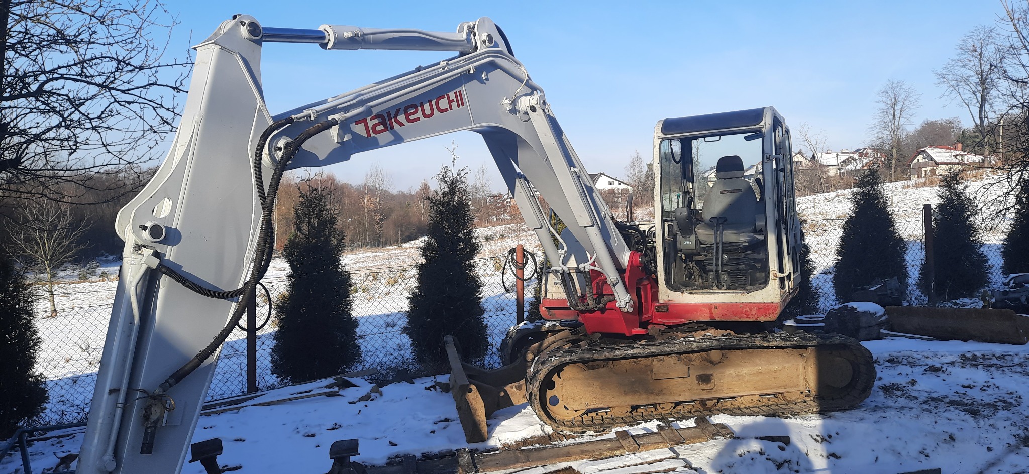 Mini koparka Takeuchi na śniegu, widoczne ślady użytkowania, w tle domy i drzewa, jasne niebo.