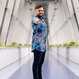 Mężczyzna w kwiecistej koszuli, stojący w nowoczesnym korytarzu, wskazuje palcem w kierunku widza, uśmiechając się.