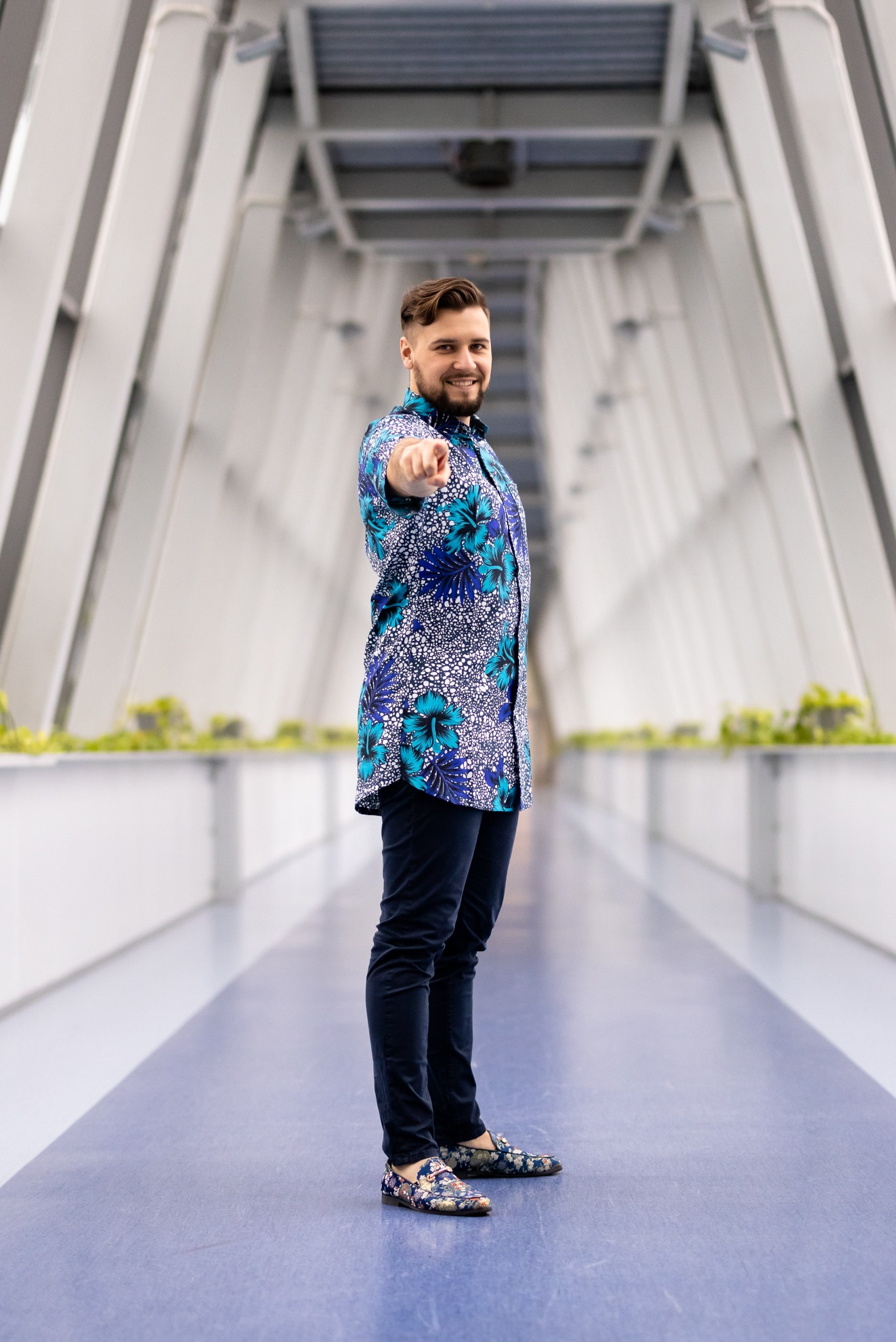 Mężczyzna w kwiecistej koszuli, stojący w nowoczesnym korytarzu, wskazuje palcem w kierunku widza, uśmiechając się.