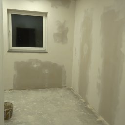 Dariusz Wiligała - Pokój w trakcie szpachlowania: ściany z widocznymi łatami gładzi, okno, sufit kasetonowy i wiadro farby w rogu. Podłoga pokryta resztkami materiałów budowlanych.