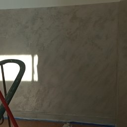 Dariusz Wiligała - Ściana w korytarzu po szpachlowaniu dekoracyjnym, widoczne ślady po taśmie malarskiej, grzejnik i fragment schodów z zieloną barierką.