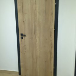 Dariusz Wiligała - Zamknięte drzwi wewnętrzne z drewnianą okleiną i czarną ościeżnicą, widoczne na tle białej ściany i podłogi z paneli. Minimalistyczny design.