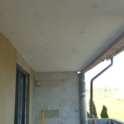 Dariusz Wiligała - Balkon w trakcie szpachlowania sufitu, widoczna drabina, poziomica i ślady po szpachli na suficie. Ściany surowe, okno z parapetem. Na podłodze resztki materiałów.