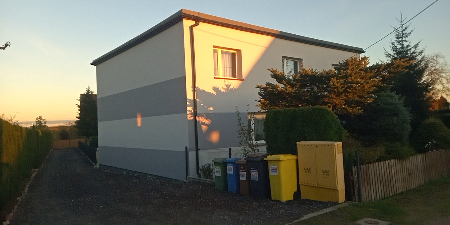 Dwukondygnacyjny dom z ocieploną elewacją w odcieniach bieli i szarości, z oknami i widocznymi koszami na śmieci. Ujęcie w ciepłym świetle zachodzącego słońca.