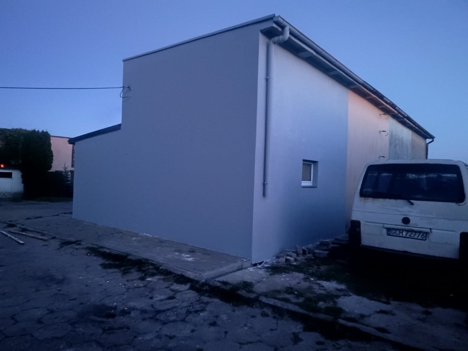 Świeżo otynkowana ściana budynku w odcieniach szarości, z widoczną rynną i małym oknem, obok zaparkowany biały bus. Ujęcie z dołu, wieczorne światło.