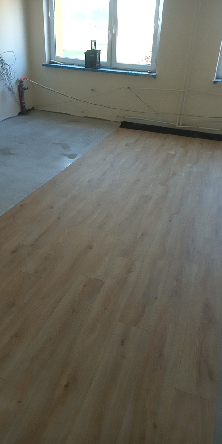 Fragment pokoju z ułożonymi panelami podłogowymi w odcieniach beżu, widoczne przygotowane podłoże i listwa przypodłogowa. Na parapecie stoi skrzynka narzędziowa.