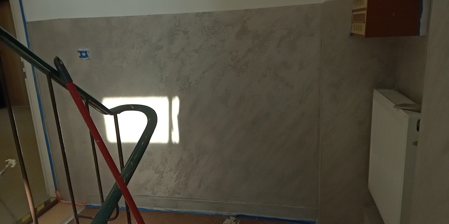 Ściana w korytarzu po szpachlowaniu dekoracyjnym, widoczne ślady po taśmie malarskiej, grzejnik i fragment schodów z zieloną barierką.