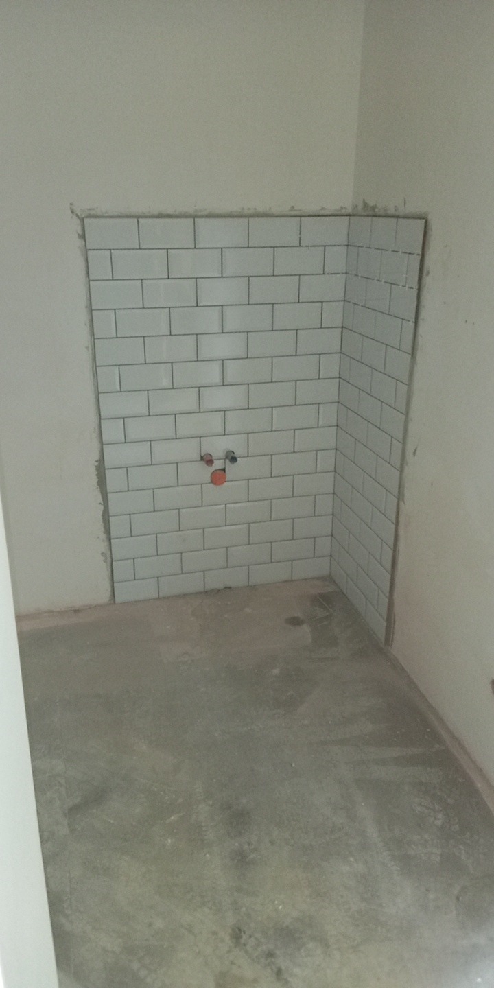Świeżo ułożone białe, prostokątne płytki na ścianie wnęki, z widocznymi przyłączami wodnymi. Surowy beton na podłodze i niedokończone ściany.