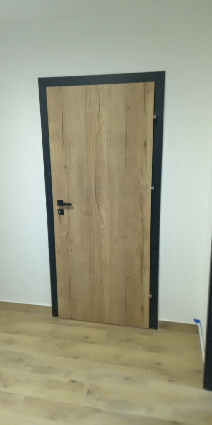 Zamknięte drzwi wewnętrzne z drewnianą okleiną i czarną ościeżnicą, widoczne na tle białej ściany i podłogi z paneli. Minimalistyczny design.