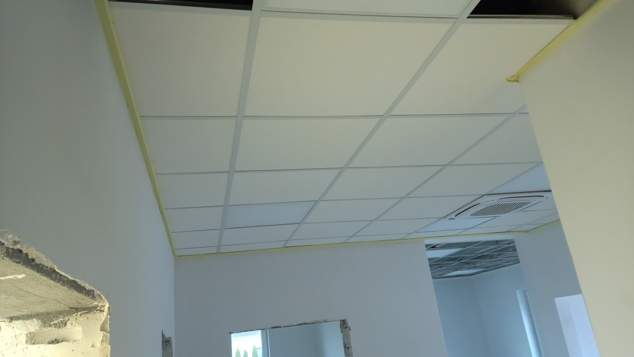 Biały sufit podwieszany kasetonowy z kratownicą, widoczny klimatyzator. Ściany pomalowane na biało, zabezpieczone taśmą malarską przy suficie. Fragment ściany w stanie surowym.
