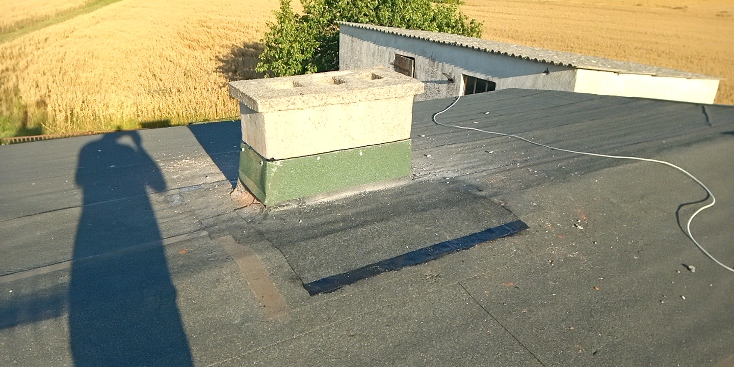 Wykończony dach pokryty papą z kominem, na tle pola i budynku gospodarczego. Widoczny cień osoby robiącej zdjęcie. Uszczelnienie wokół komina.