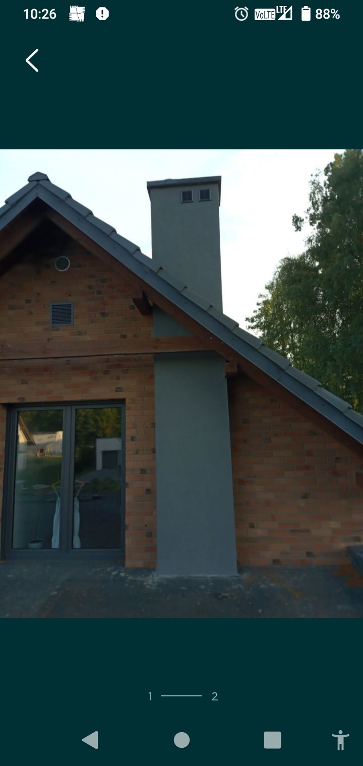 Wykończony komin w szarym kolorze, przylegający do ściany budynku z cegły w Kamieniu Krajeńskim. Widoczny fragment dachu i okno.