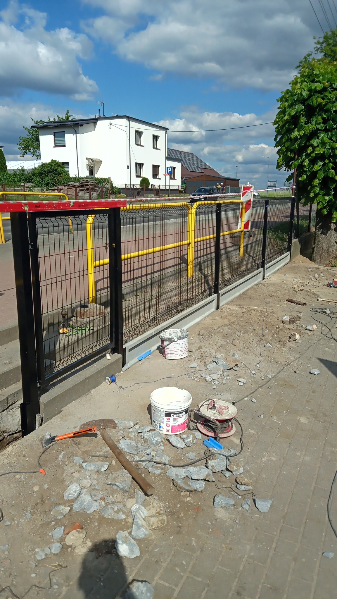 Nowoczesne, czarne ogrodzenie panelowe z żółtymi elementami, osadzone na betonowej podmurówce. Widoczne narzędzia i materiały budowlane sugerują etap montażu.