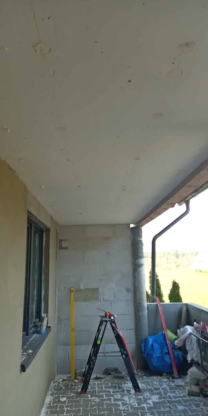 Balkon w trakcie szpachlowania sufitu, widoczna drabina, poziomica i ślady po szpachli na suficie. Ściany surowe, okno z parapetem. Na podłodze resztki materiałów.