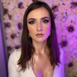 Make Up Studio Amelia Mazurowska - Portret kobiety z profesjonalnie wykonanym smokey eye i błyszczącymi akcentami w kącikach oczu, na tle ściany z róż w odcieniach fioletu i różu, z delikatnymi kolczykami.