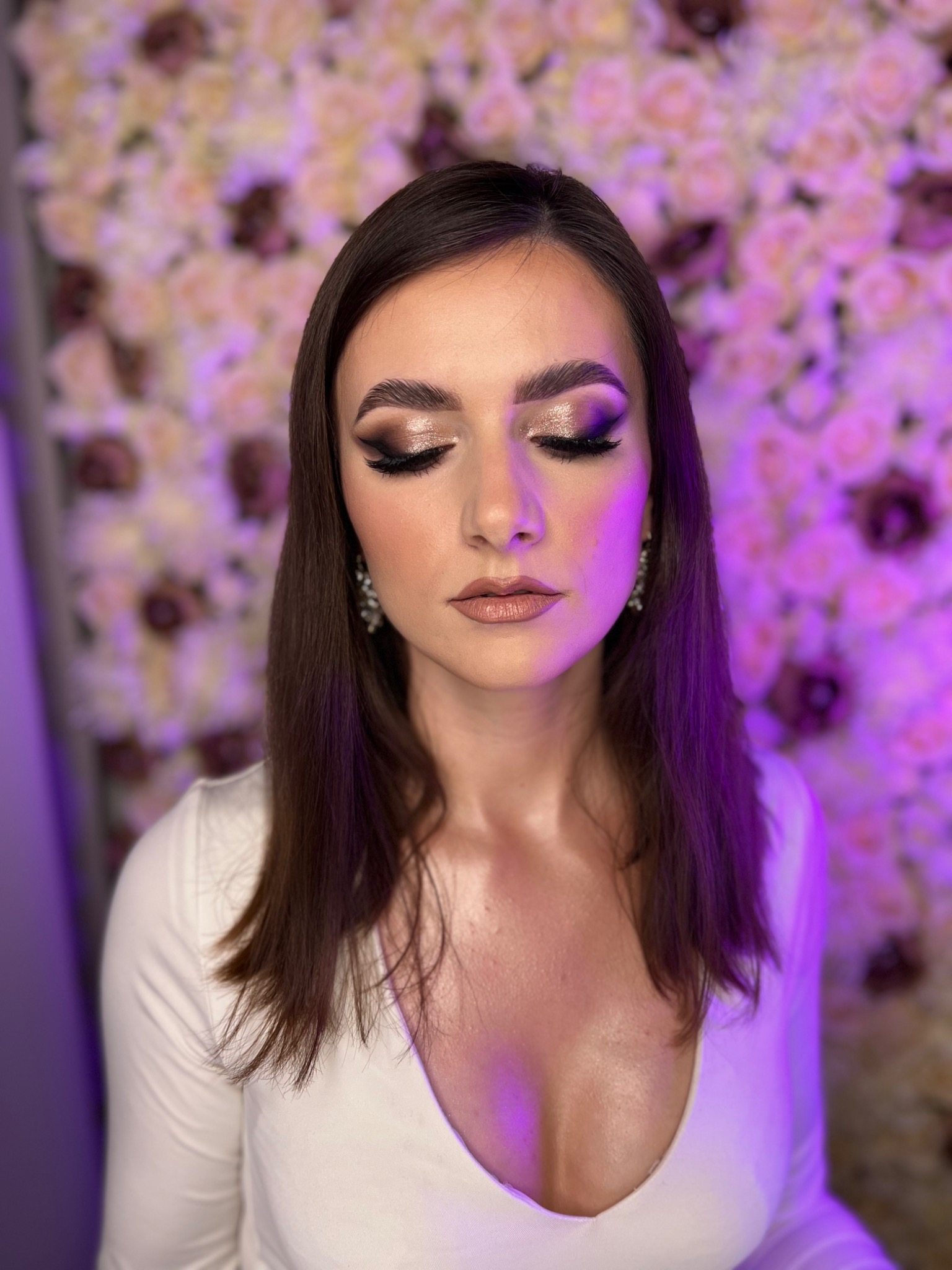 Portret kobiety z zamkniętymi oczami, prezentującej wieczorowy look z błyszczącym cieniem, podkreślonym eyelinerem i subtelnym konturowaniem, na tle ściany z kwiatów w odcieniach różu i fioletu.