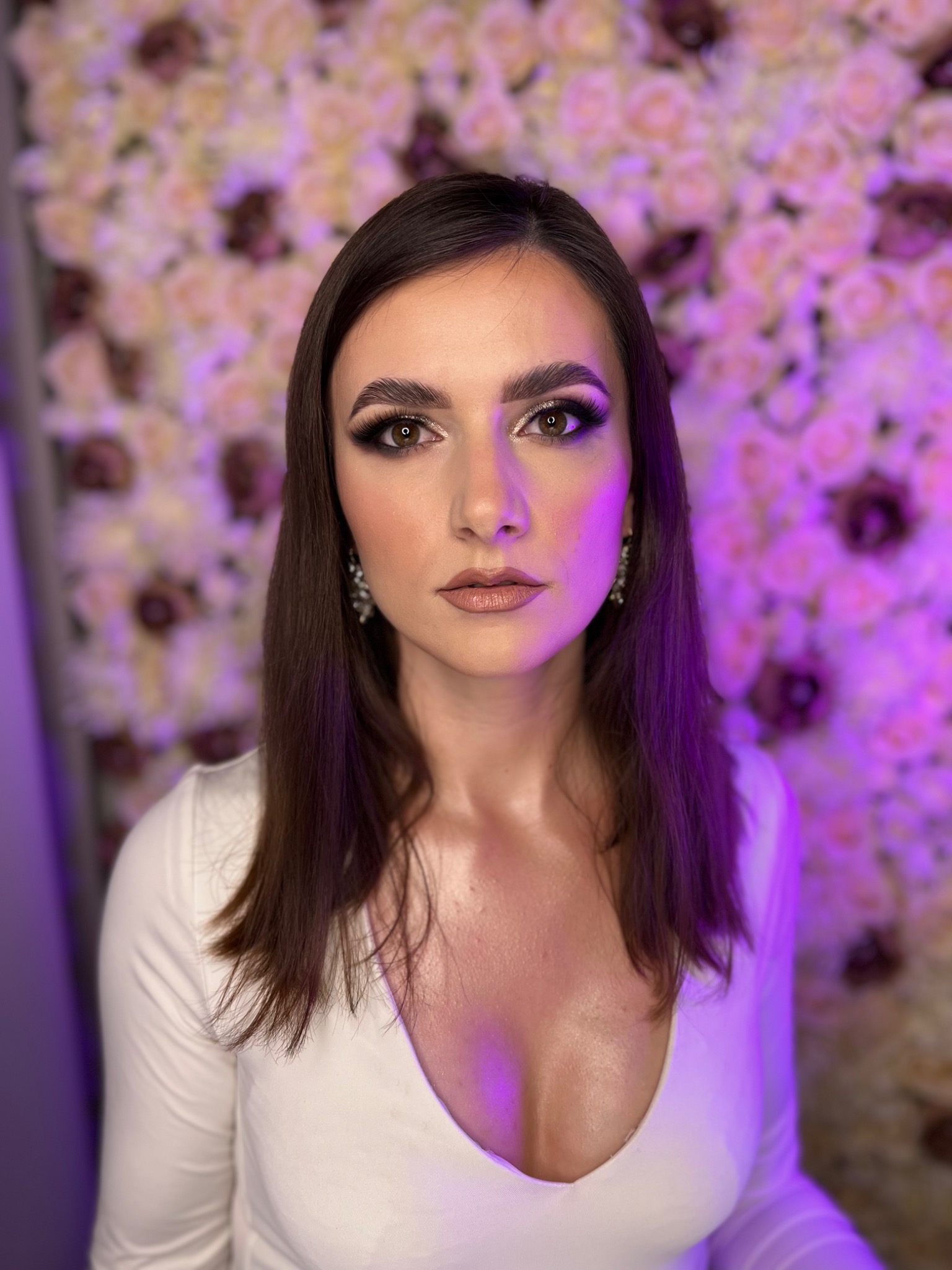 Portret kobiety z profesjonalnie wykonanym smokey eye i błyszczącymi akcentami w kącikach oczu, na tle ściany z róż w odcieniach fioletu i różu, z delikatnymi kolczykami.