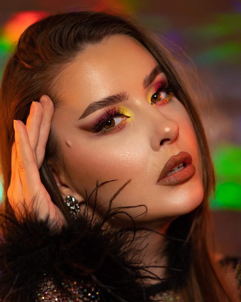 Portret kobiety z intensywnym, kolorowym wizażem oczu w odcieniach żółci i różu, podkreślonym eyelinerem, oraz błyszczącymi, brązowymi ustami; dłoń przy skroni, błyszczące kolczyki, tło z efektem...