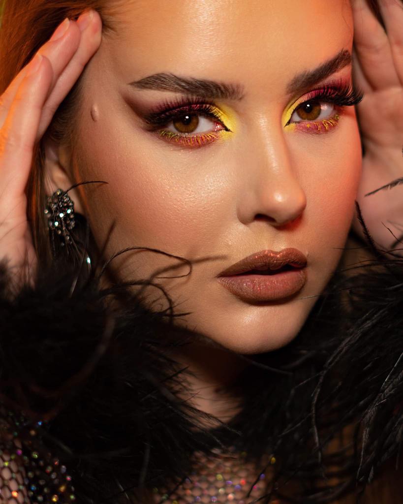 Portret kobiety z intensywnym makijażem oczu w odcieniach żółci, różu i bordo, z wyraźnie podkreślonymi brwiami i ustami w kolorze nude; stylizacja uzupełniona o czarne pióra.