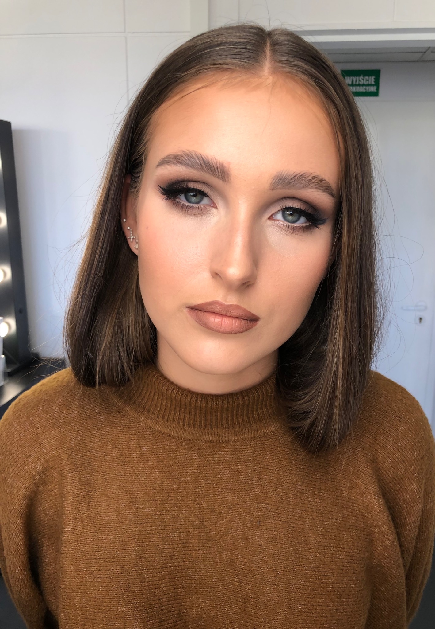 Portret kobiety z profesjonalnym, wieczorowym w odcieniach brązu, z wyraźnie zaznaczonymi liniami rzęs i ustami w kolorze nude, na tle białej ściany i czarnej szafki z oświetleniem.