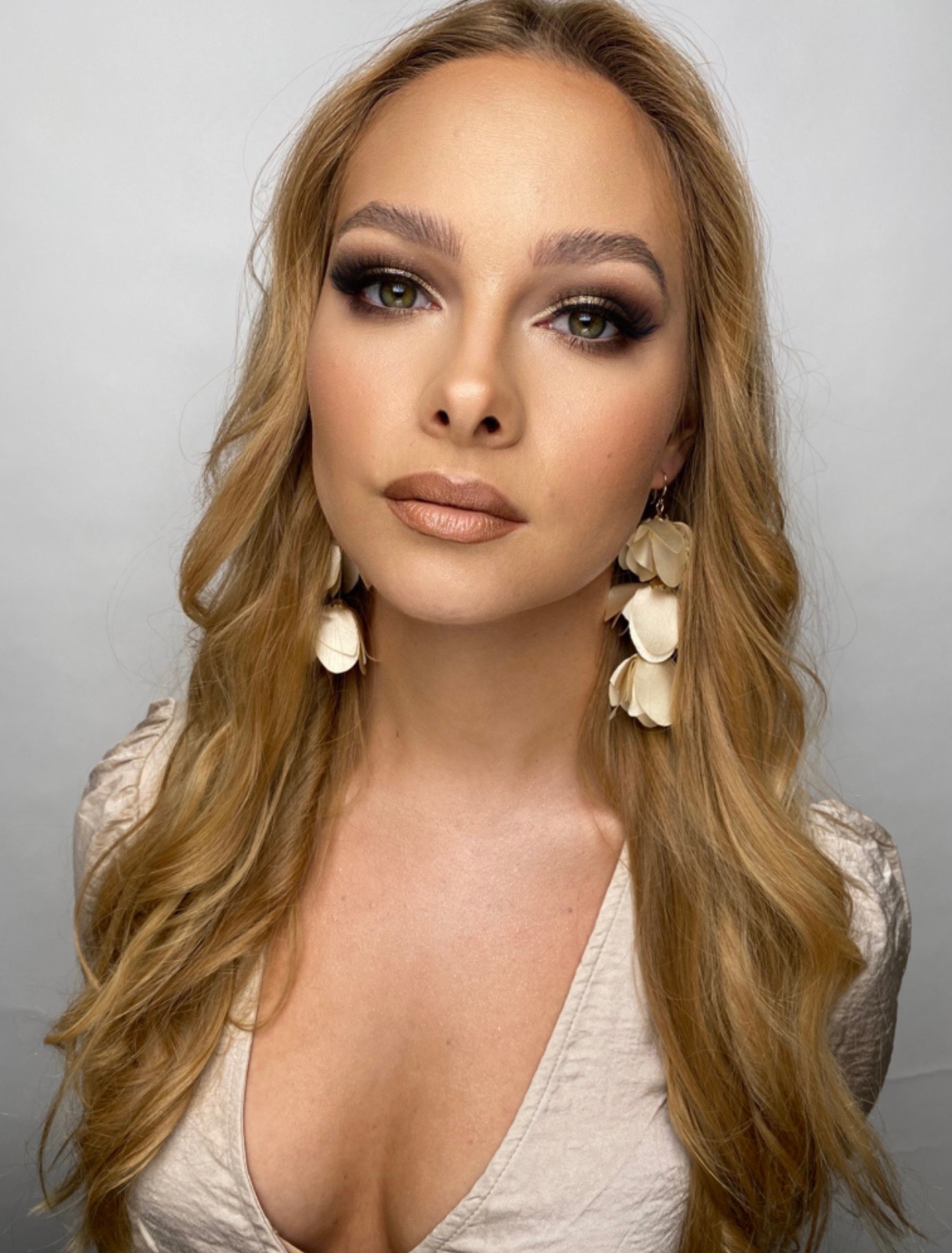 Portret kobiety z profesjonalnie wykonanym, złotym makijażem oczu i ust, długimi blond włosami i dużymi, kremowymi kolczykami w kształcie kwiatów.
