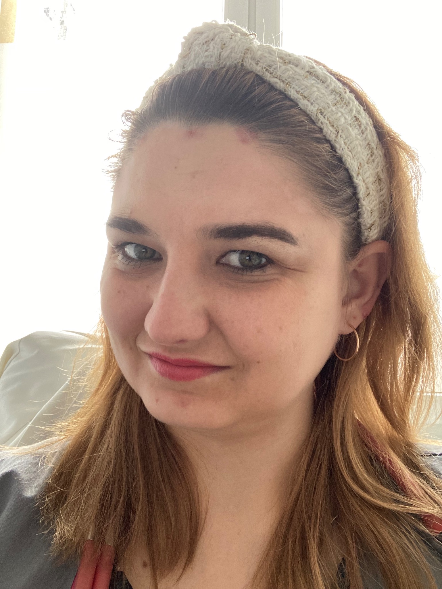 Portret uśmiechniętej kobiety z blond włosami i beżową opaską na głowie, złote kolczyki koła, delikatny makijaż.