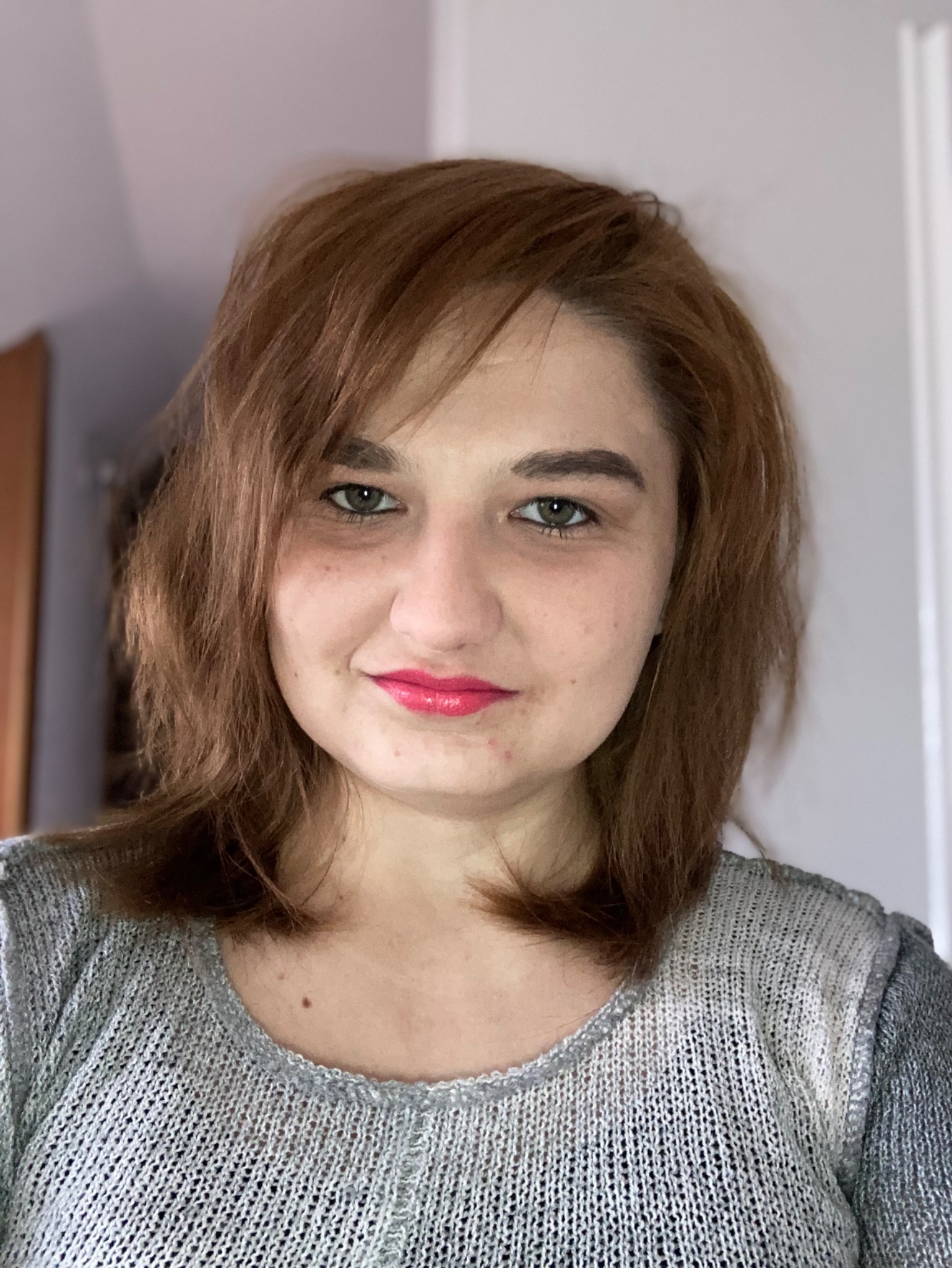 Portret kobiety z brązowymi włosami, uśmiechającej się, z różową szminką, w szarym swetrze z dzianiny, na rozmytym tle ściany i drzwi.