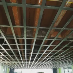 Stelaż sufitu podwieszanego z widoczną starą konstrukcją drewnianą i przewodami elektrycznymi w tle.