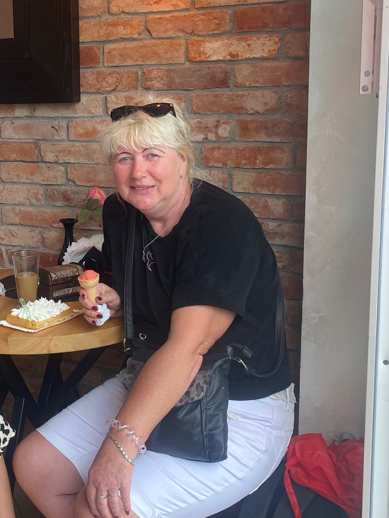 Uśmiechnięta kobieta z blond włosami i okularami na głowie, siedzi przy stoliku z gofrem z bitą śmietaną i lodami w wafelku na tle ceglanej ściany.