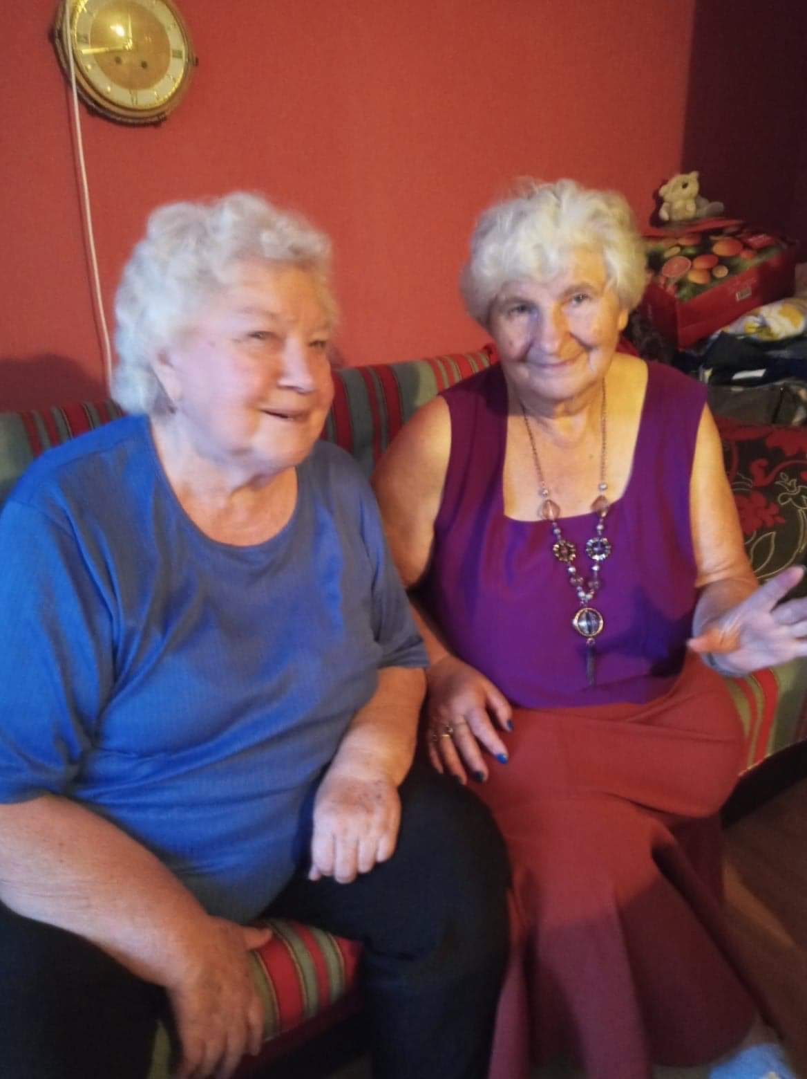 Dwie uśmiechnięte starsze kobiety siedzą na kanapie na tle czerwonej ściany z wiszącym zegarem i pudełkiem z pluszowym misiem. Kobieta po lewej w niebieskiej koszulce patrzy na drugą kobietę...