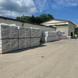 Grupa Gamma - Skład budowlany: palety wełny mineralnej Petralana i worki z zaprawą na kostce brukowej, budynek w tle pod błękitnym niebem.