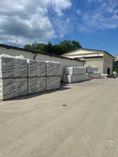Skład budowlany: palety wełny mineralnej Petralana i worki z zaprawą na kostce brukowej, budynek w tle pod błękitnym niebem.