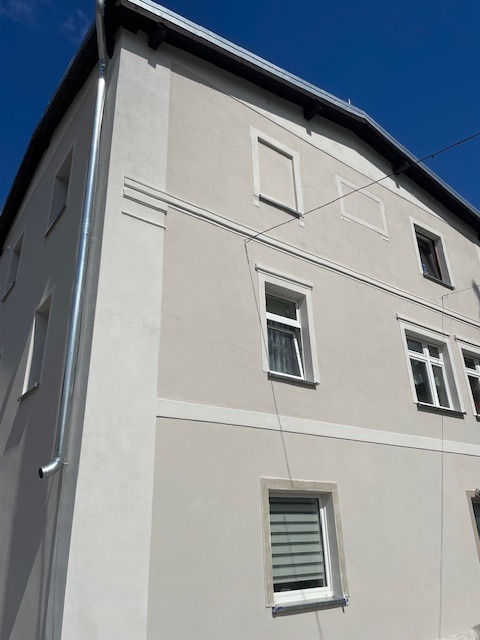 Elewacja budynku po remoncie z nowym tynkiem w odcieniach szarości, białe obramowania okien i rynna spustowa po lewej stronie. Błękitne niebo w tle.