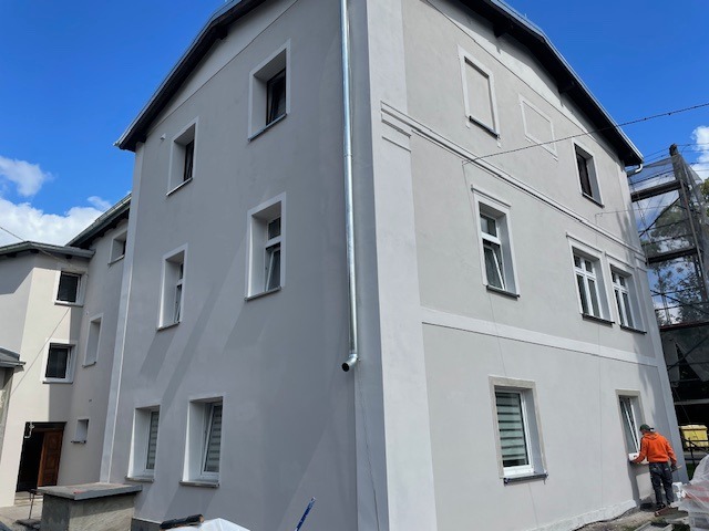 Odnowiona elewacja budynku wielorodzinnego w odcieniach szarości, z białymi obramowaniami okien. Pracownik maluje dolną część fasady. Widoczne rusztowanie.