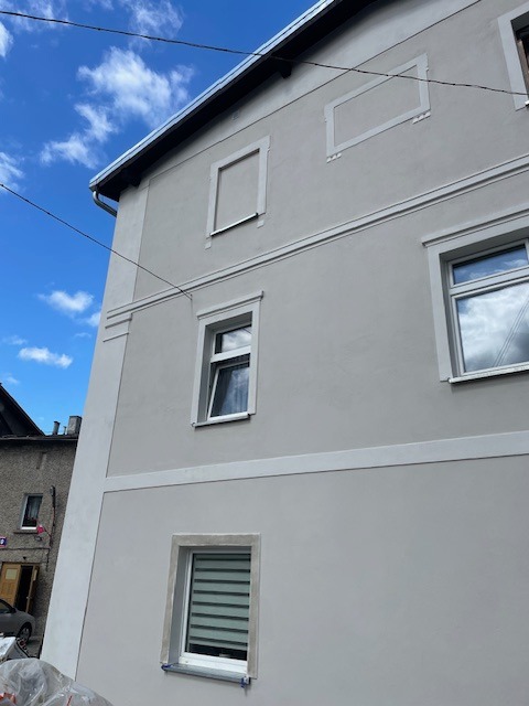 Elewacja budynku po tynkowaniu maszynowym, widok z dołu na fragment ściany z oknami i dekoracyjnymi obramowaniami. Jasne, neutralne kolory.