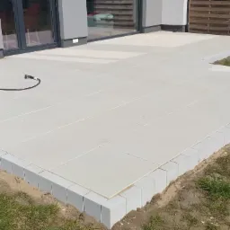 Jasnoszary taras z kostki brukowej ułożony przy nowoczesnym domu. Widoczny fragment trawnika i narzędzie pracy - żółty pojemnik.