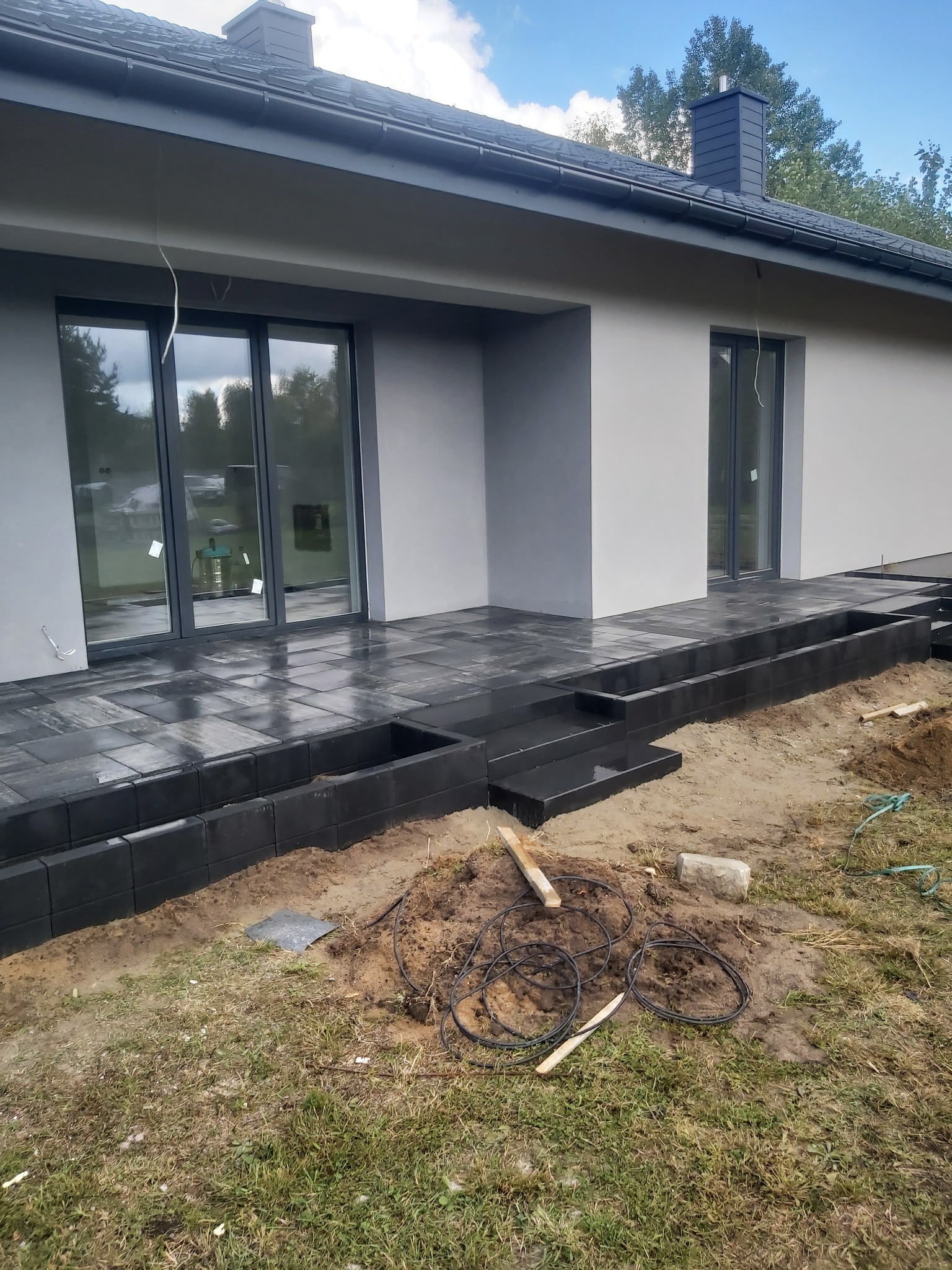 Nowoczesny taras z ciemnoszarej kostki brukowej przy domu z szarą elewacją, widoczne schody i rabaty, teren wokół w trakcie prac ziemnych, zwinięte kable na pierwszym planie.