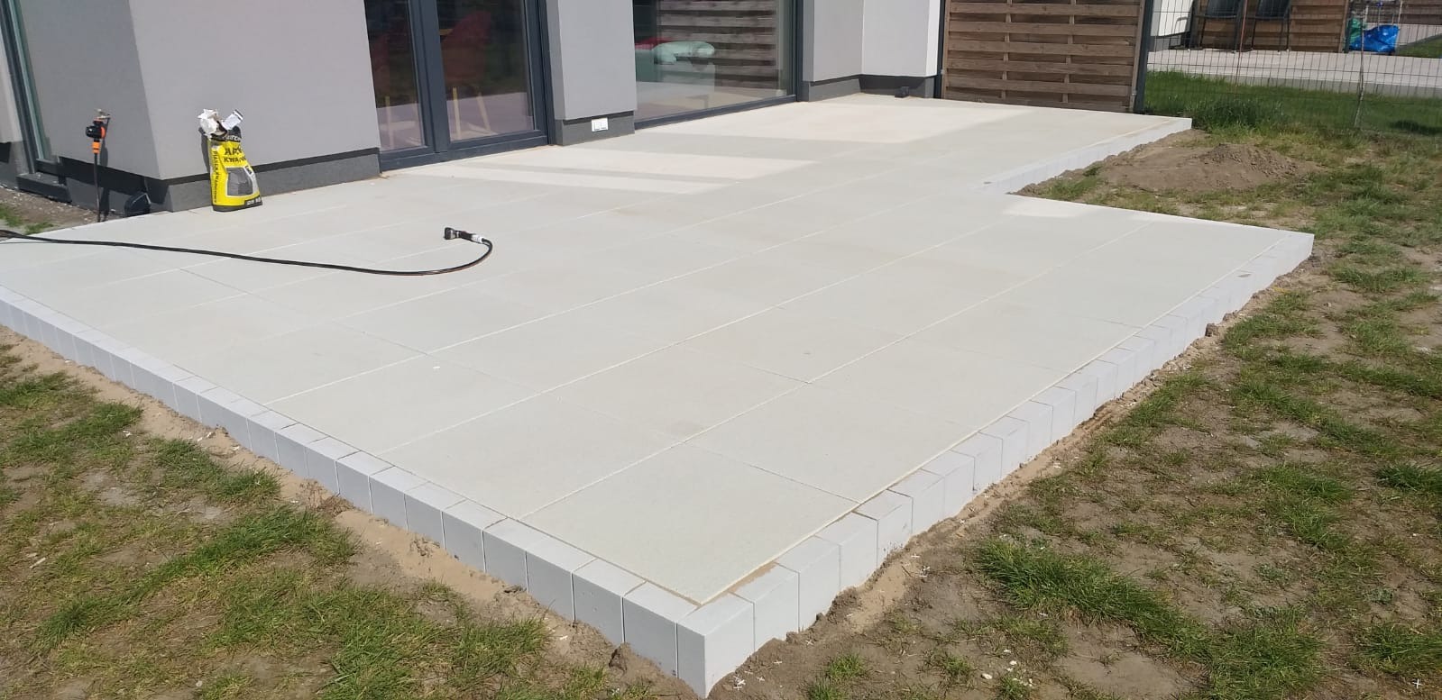 Jasnoszary taras z kostki brukowej ułożony przy nowoczesnym domu. Widoczny fragment trawnika i narzędzie pracy - żółty pojemnik.