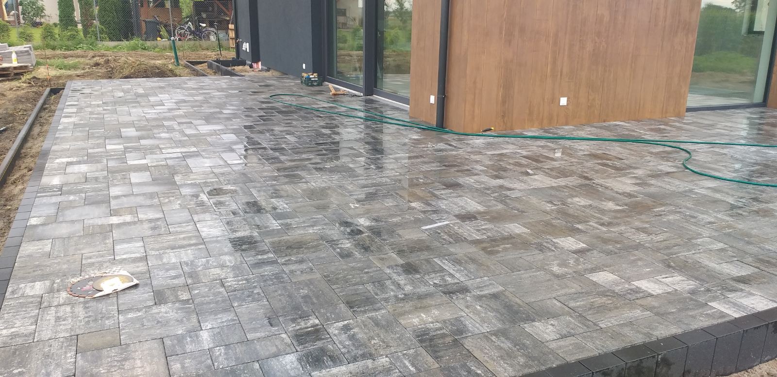 Ułożona, szara kostka brukowa przed nowoczesnym budynkiem z drewnianą elewacją. Widoczny fragment ogrodu i narzędzia. Kostka mokra, odbija światło.