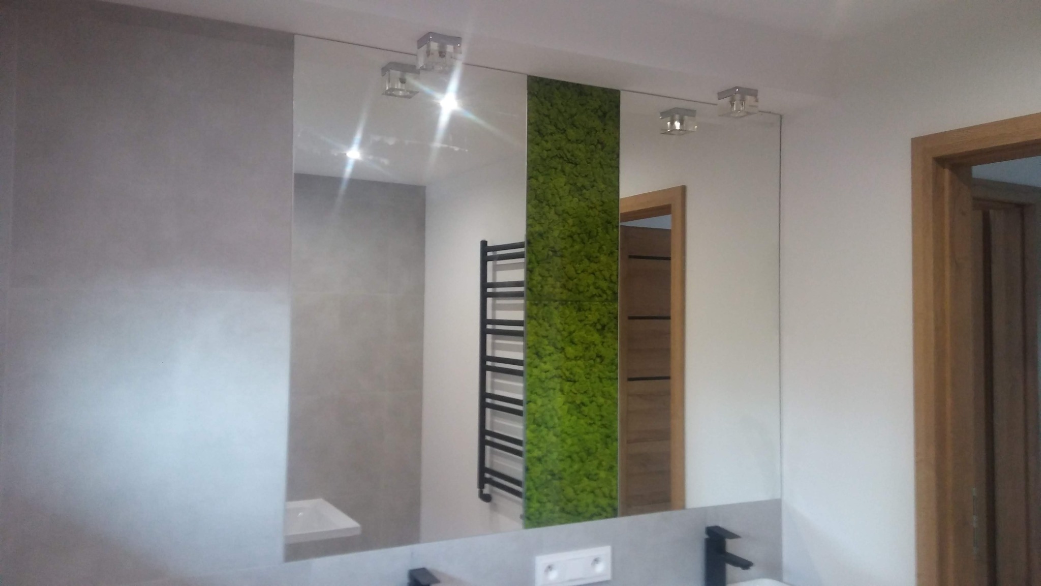 Nowoczesna łazienka z dużym lustrem, pionowym panelem z mchu, czarnym grzejnikiem drabinkowym i minimalistyczną armaturą.