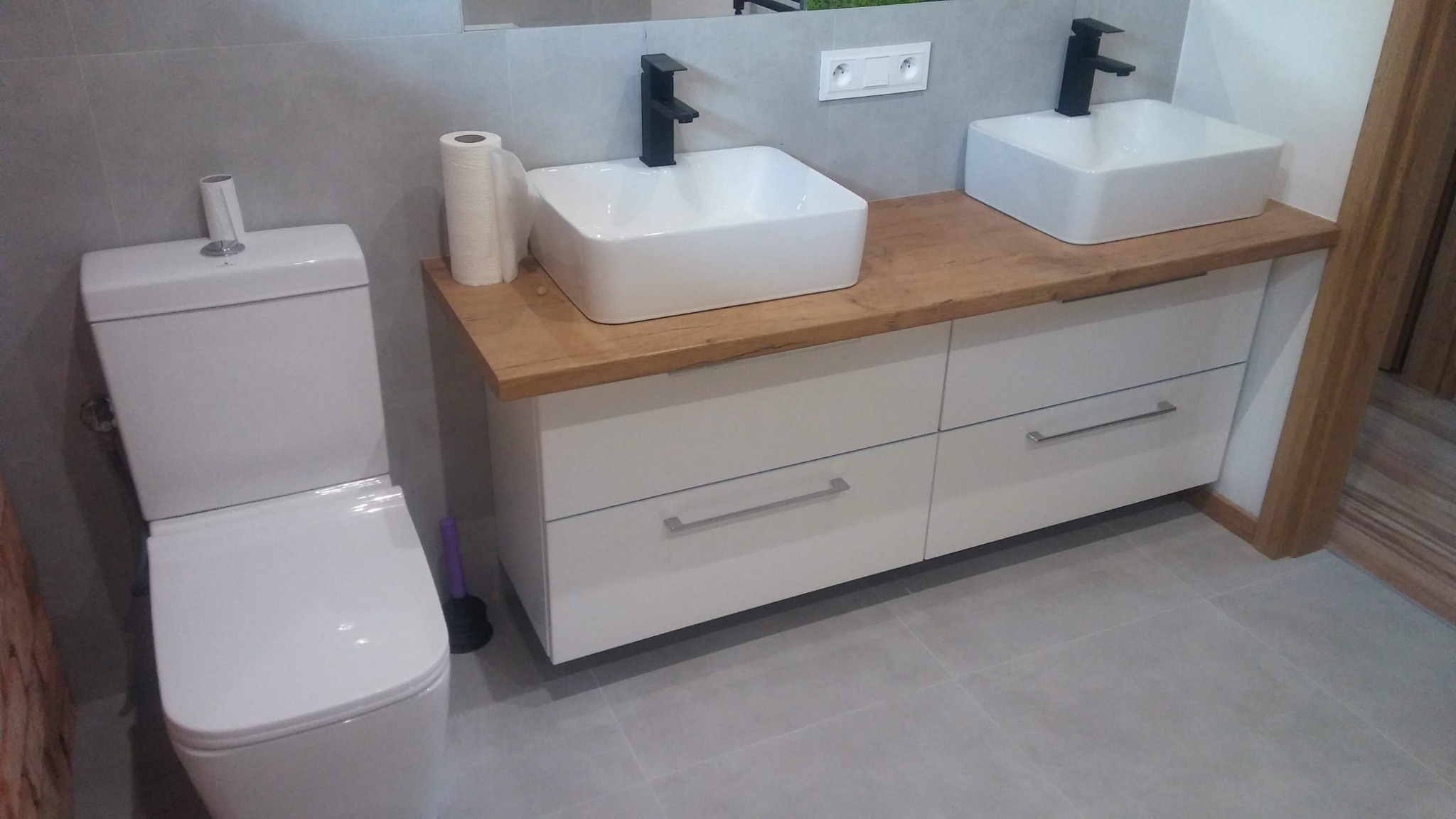 Nowoczesna łazienka z dwoma białymi umywalkami nablatowymi z czarnymi bateriami, drewnianym blatem, białą szafką z szufladami i toaletą w tle, uzupełniona o rolkę papieru toaletowego i fioletową...