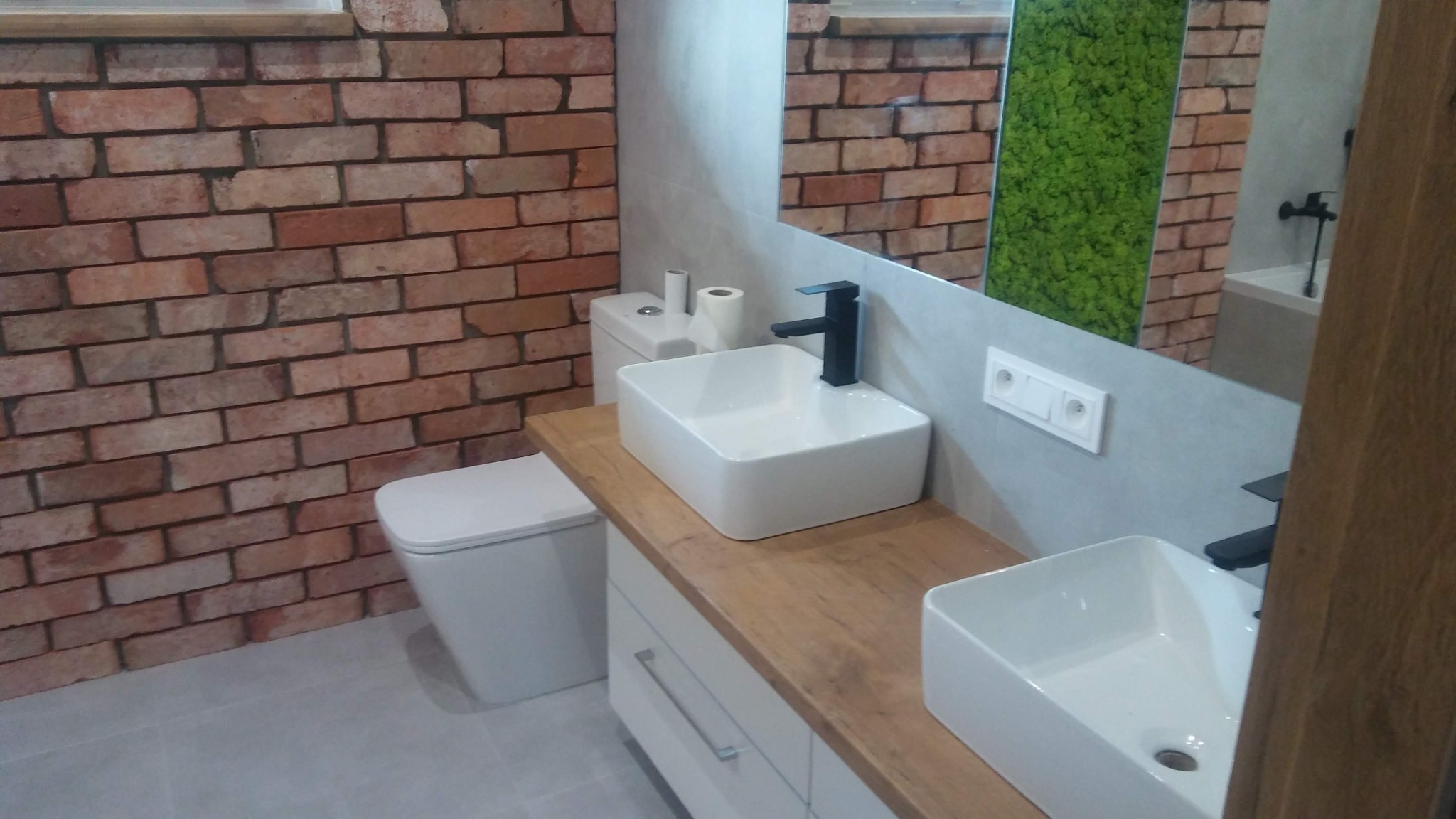 Nowoczesna łazienka z umywalkami nablatowymi, bateriami w kolorze czarnym, ceglaną ścianą i panelem z mchu w lustrze.